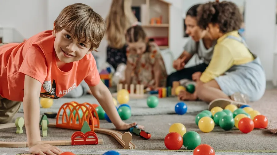 Skupina dětí si hraje v herně s barevnými hračkami a míčky, v popředí chlapec ve světle červeném tričku